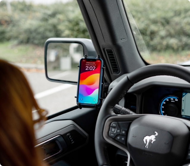 A driver’s-side view of a Ford Bronco interior with a smartphone secured in a RAM X-Grip mount attached to the A-pillar. The phone is clearly visible and accessible above the steering wheel, providing safe hands-free usage for driving or trail navigation.