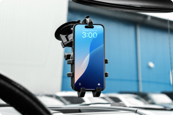 A smartphone is securely held in a RAM Quick-Grip holder with a twist-lock suction cup base, mounted to the inside of a vehicle’s windshield. The setup positions the phone centrally for optimal visibility and hands-free access, with a blurred background of parked vehicles and a blue building.