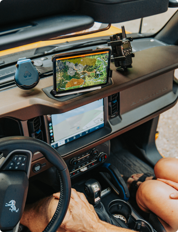 Top-down view of a Ford Bronco dashboard equipped with a full navigation and communication setup. A rugged tablet displaying topographic maps is secured with a RAM Mount, next to a mounted two-way radio and a heart-shaped puck charger. The setup supports hands-free GPS use and radio communication while exploring off-road.