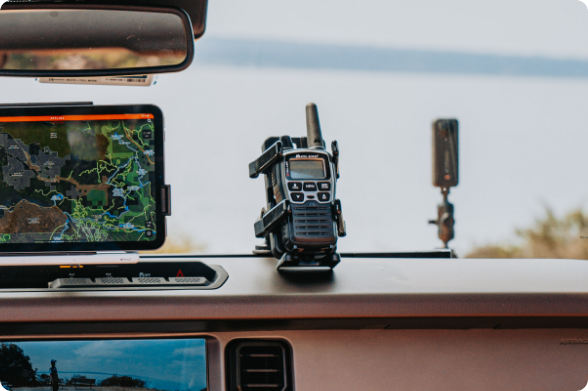A dual-device dashboard mounting setup featuring a RAM Mount tablet holder displaying a GPS topo map and a two-way radio mounted beside it. Both devices are secured to the dash of an off-road vehicle with Lake or ocean scenery visible through the windshield, illustrating the system’s use in remote navigation and communication.