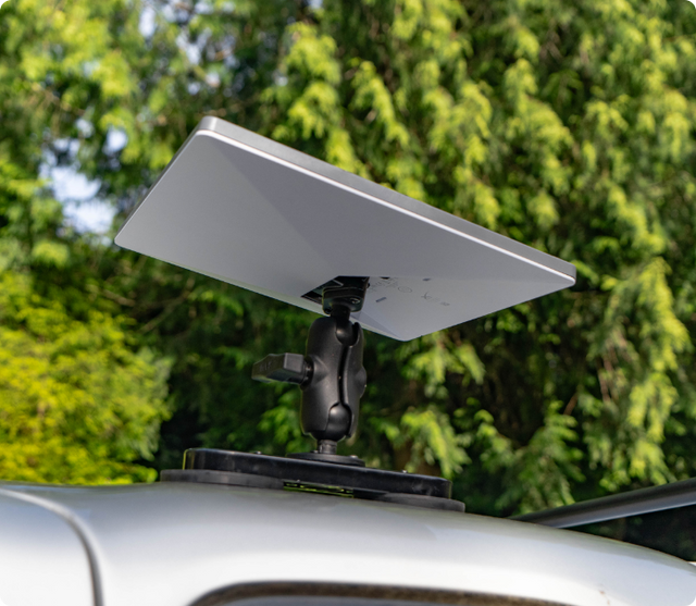 A Starlink Mini satellite dish installed on the roof of a vehicle using a RAM Mount ball and socket system. The mount is secured to a flat base, providing portable, vehicle-based internet connectivity in remote areas, with a lush green forest in the background.
