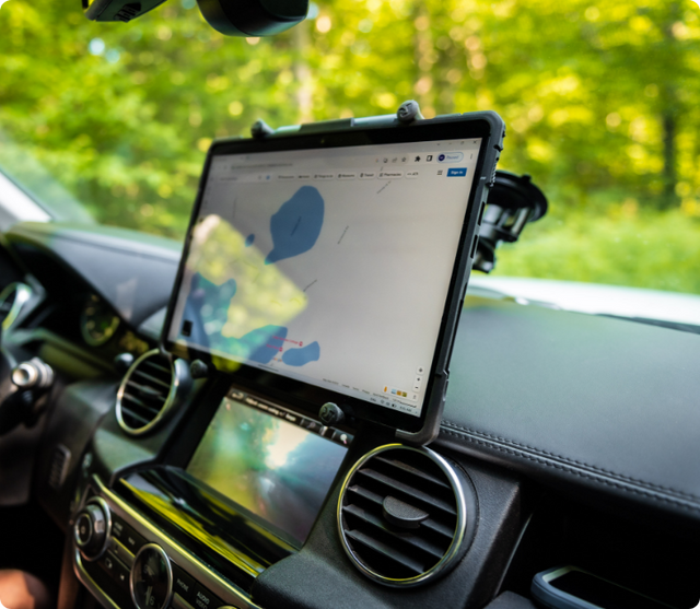 Interior of an off-road SUV with a rugged tablet mounted front and center above the infotainment display using a RAM Mount system. The tablet displays a mapping/navigation app and is held securely between the dashboard air vents with a side-mounted RAM arm, ideal for route planning on forested trails.